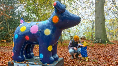 A man crouched down next to a toddler, both in woolly hats, looking at a sculpture of a dark blue Snowdog painted with large brightly coloured circles resembling lights.